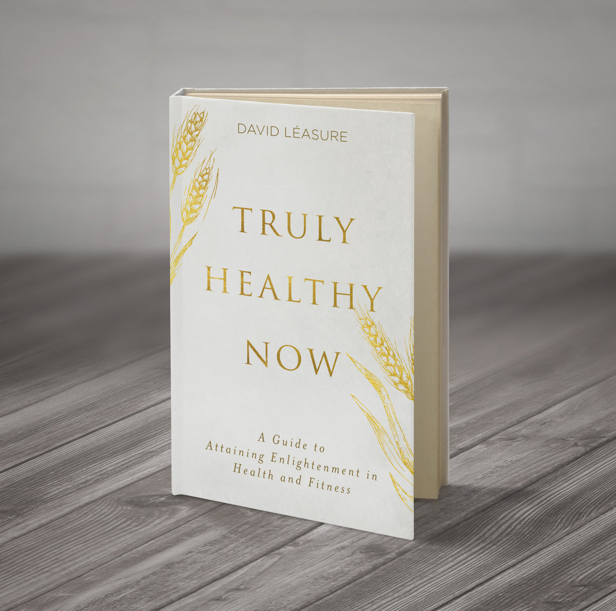 Truly Healthy Now paperback book standing upright on a wood surface — gold title text and wheat illustrations on white cover, subtitle: A Guide to Attaining Enlightenment in Health and Fitness